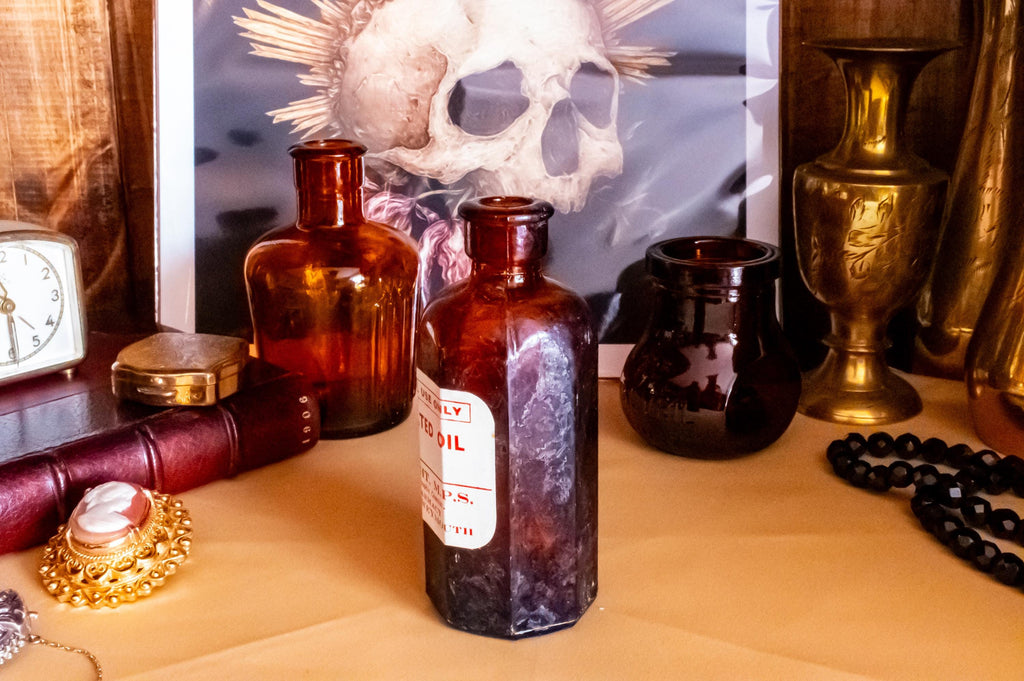 Three vintage amber glass apothecary bottles in varying shapes and sizes, one retaining a vintage label, displayed near a brass vase and skull artwork.