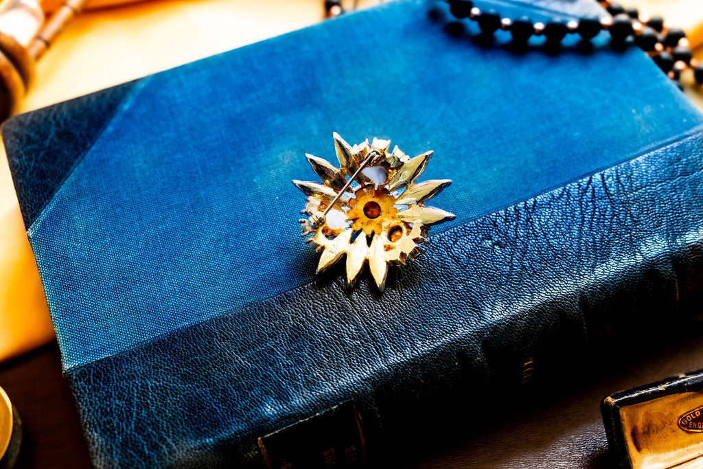 Vintage gold-tone brooch shaped like a flower with multi-layered faceted blue crystals and rhinestones, resting on a blue book.