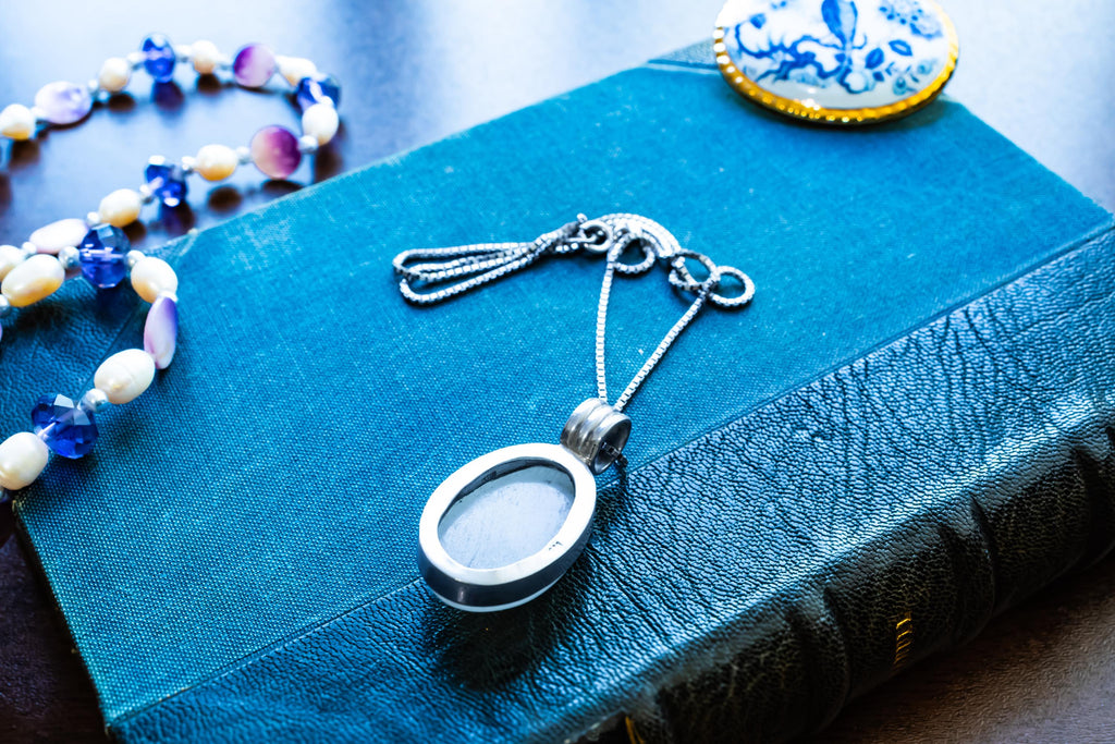 Large oval cabochon luminous moonstone pendant set in 925 sterling silver on a heavy box chain, displayed on a blue vintage book.