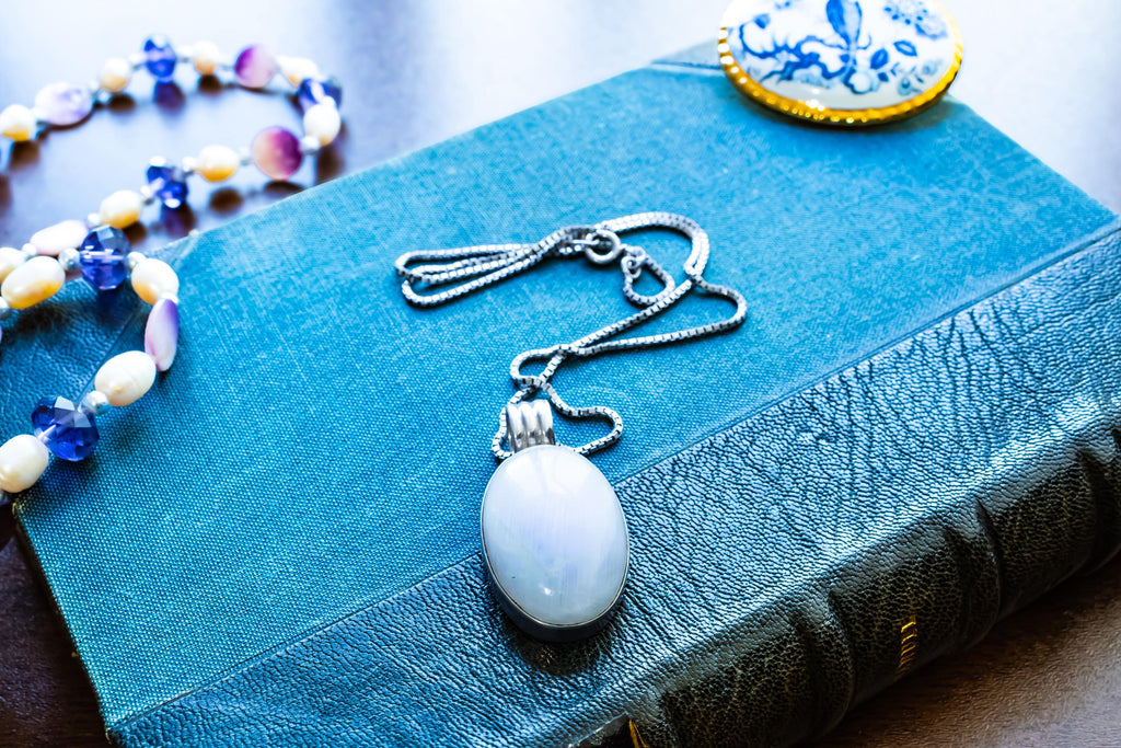 Large oval cabochon luminous moonstone pendant set in 925 sterling silver on a heavy box chain, displayed on a blue vintage book.
