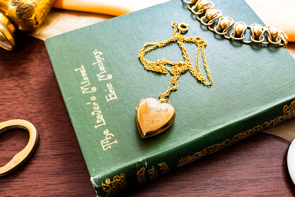 An antique 9ct gold back and front heart-shaped locket featuring a delicate hand-engraved swallow, resting romantically on a green vintage book titled "The Lover's Missal".