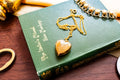 An antique 9ct gold back and front heart-shaped locket featuring a delicate hand-engraved swallow, resting romantically on a green vintage book titled "The Lover's Missal".