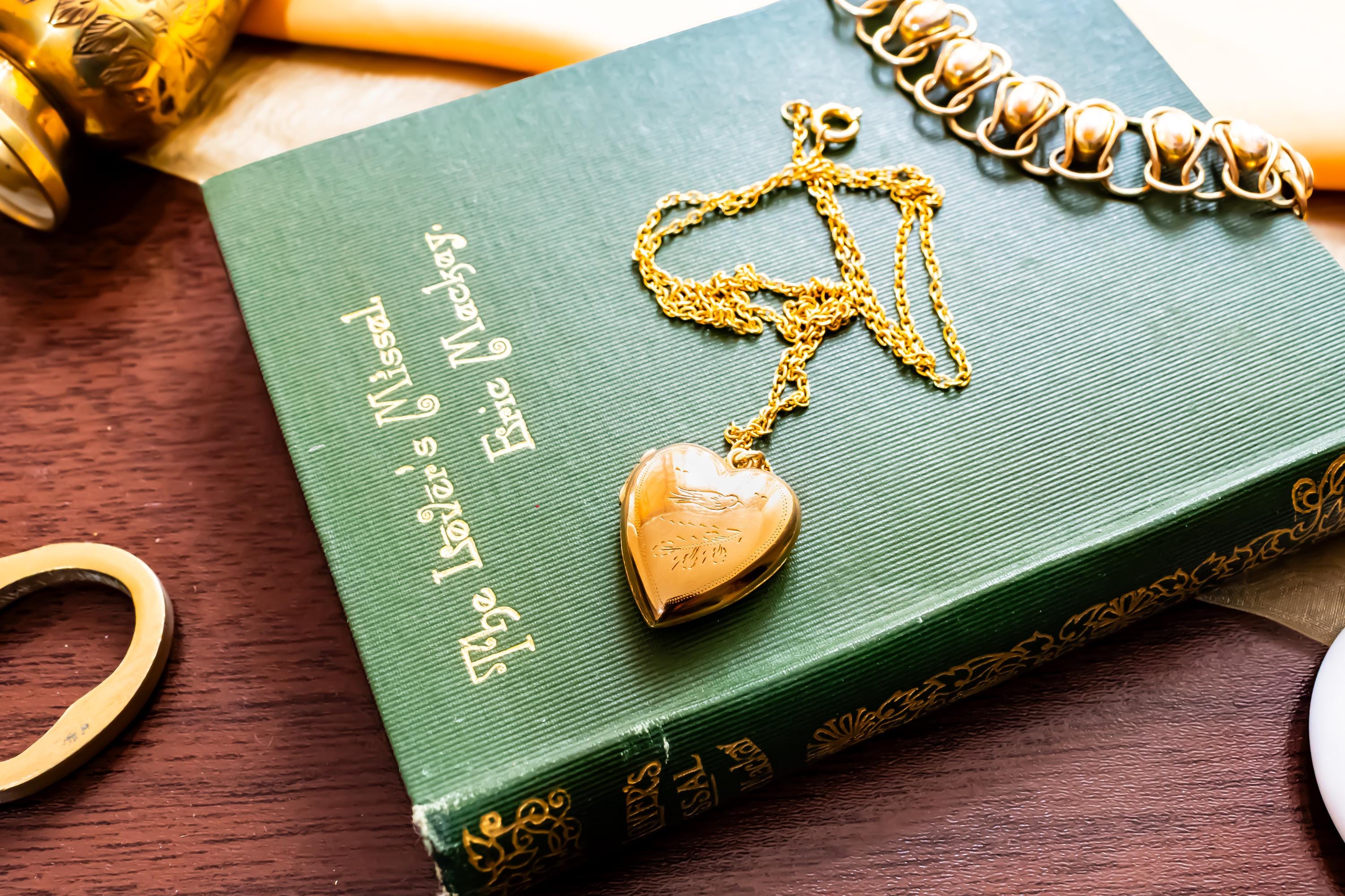 An antique 9ct gold back and front heart-shaped locket featuring a delicate hand-engraved swallow, resting romantically on a green vintage book titled "The Lover's Missal".