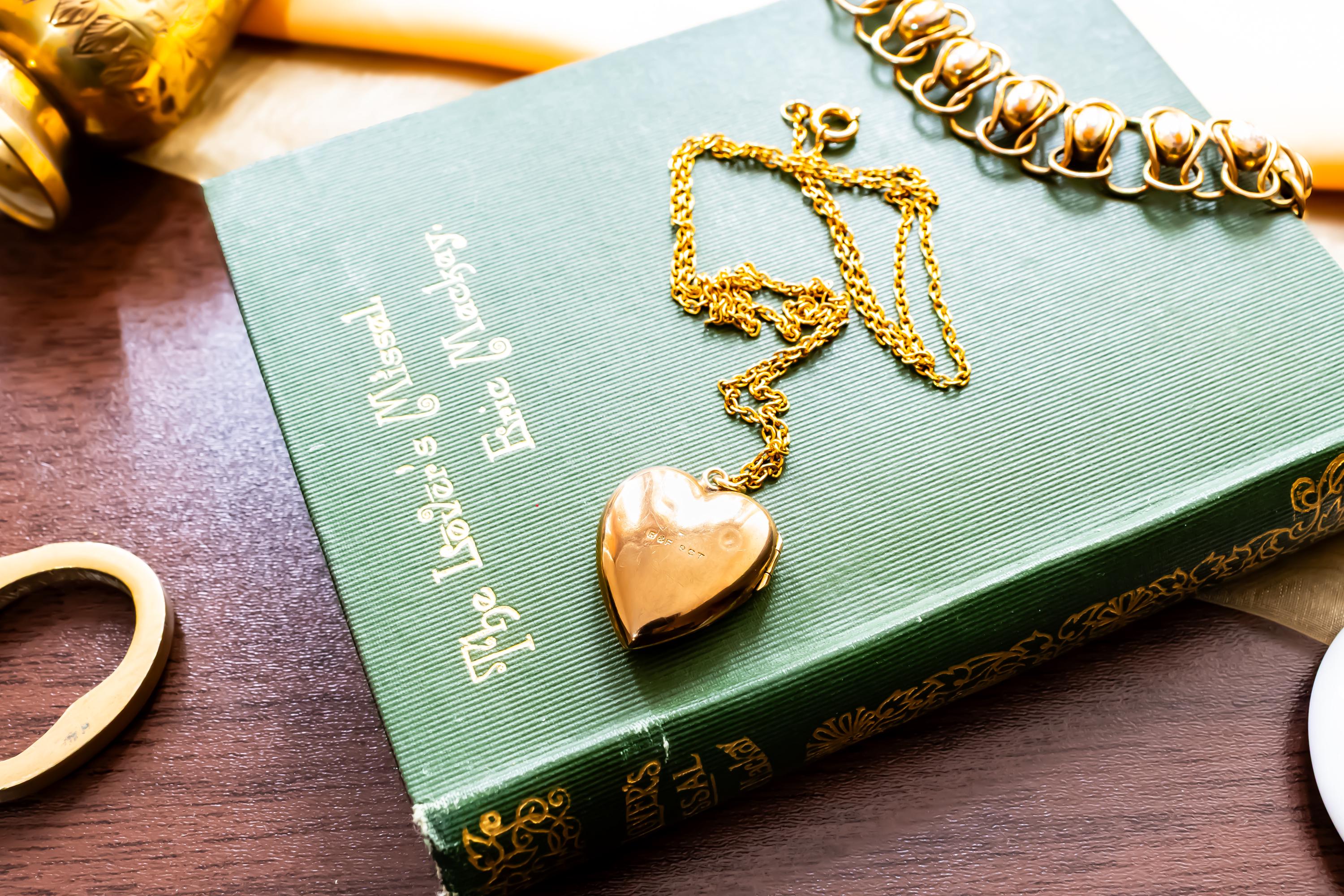 An antique 9ct gold back and front heart-shaped locket featuring a delicate hand-engraved swallow, resting romantically on a green vintage book titled "The Lover's Missal".