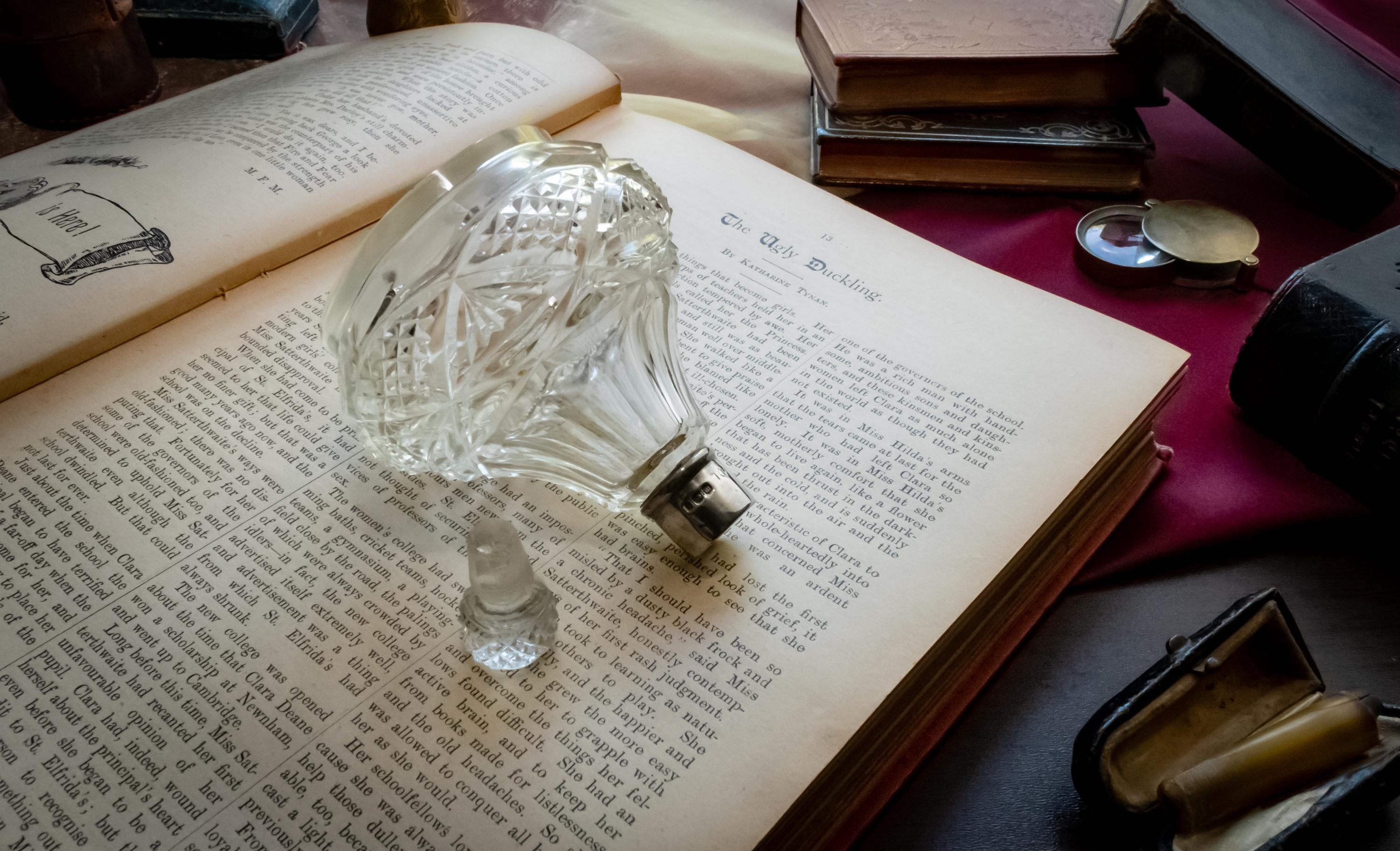 An antique heavy cut glass scent bottle with a sterling silver collar and crystal stopper, sitting on an open vintage book surrounded by dark academia props