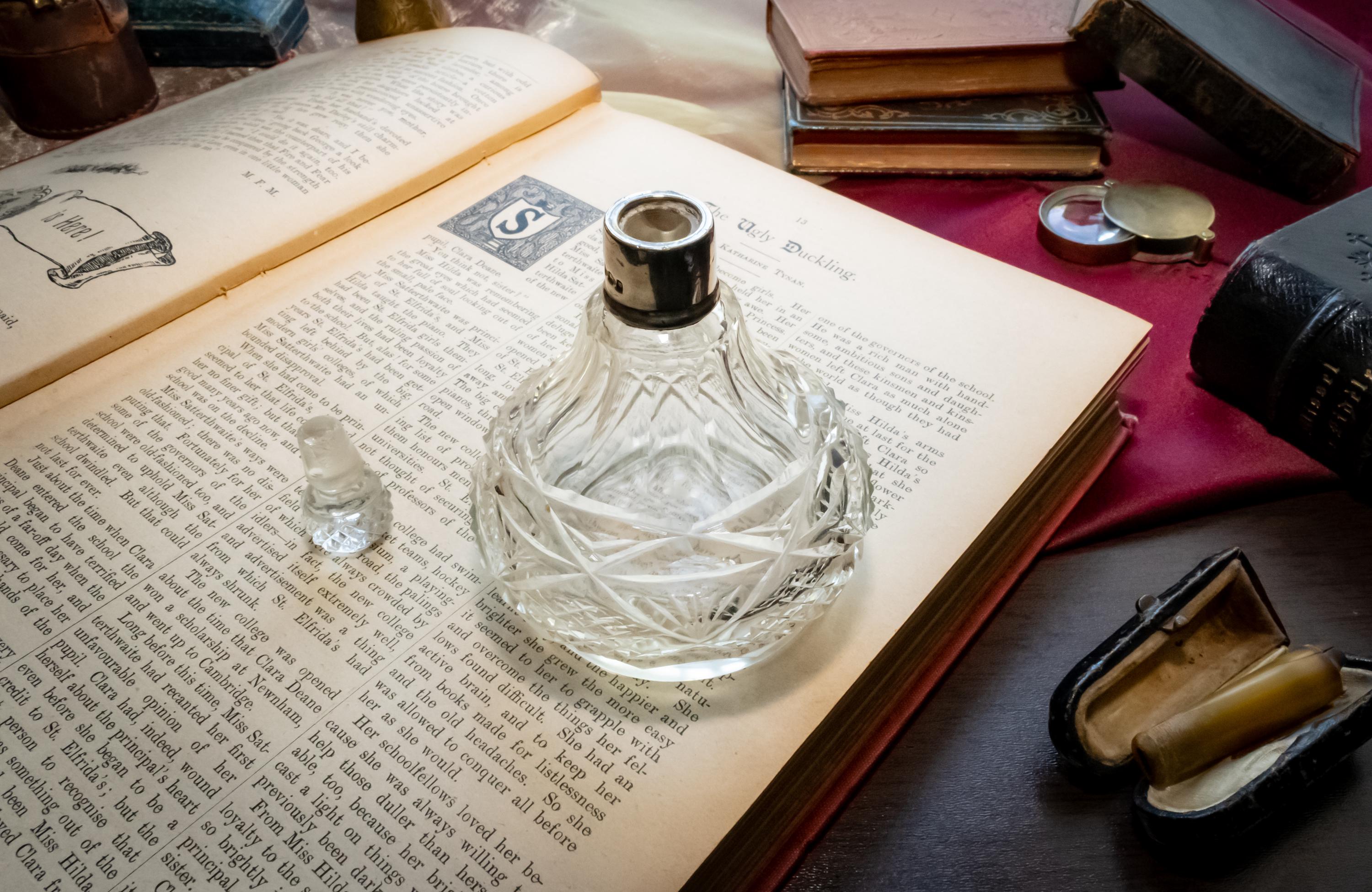 An antique heavy cut glass scent bottle with a sterling silver collar and crystal stopper, sitting on an open vintage book surrounded by dark academia props
