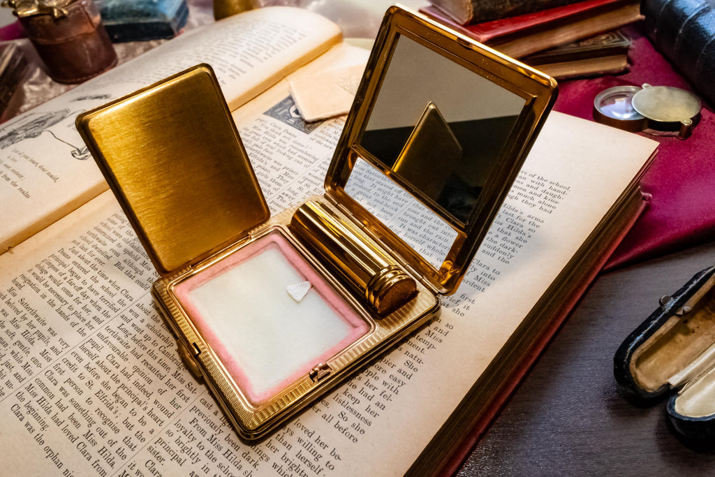 A gleaming gold vintage Kigu of London rectangular compact with a basketweave pattern and integrated lipstick holder, resting on an open antique book.