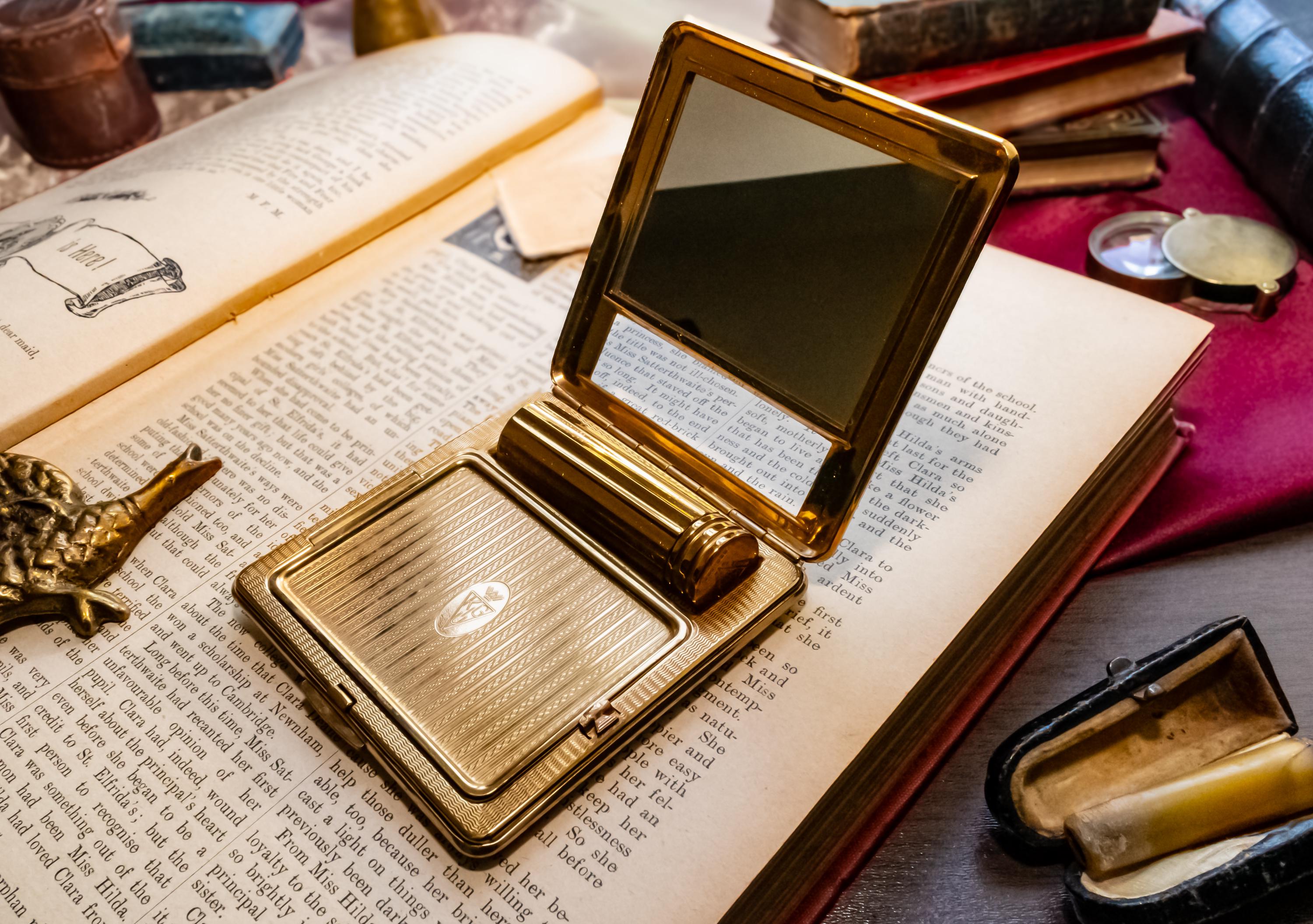A gleaming gold vintage Kigu of London rectangular compact with a basketweave pattern and integrated lipstick holder, resting on an open antique book.