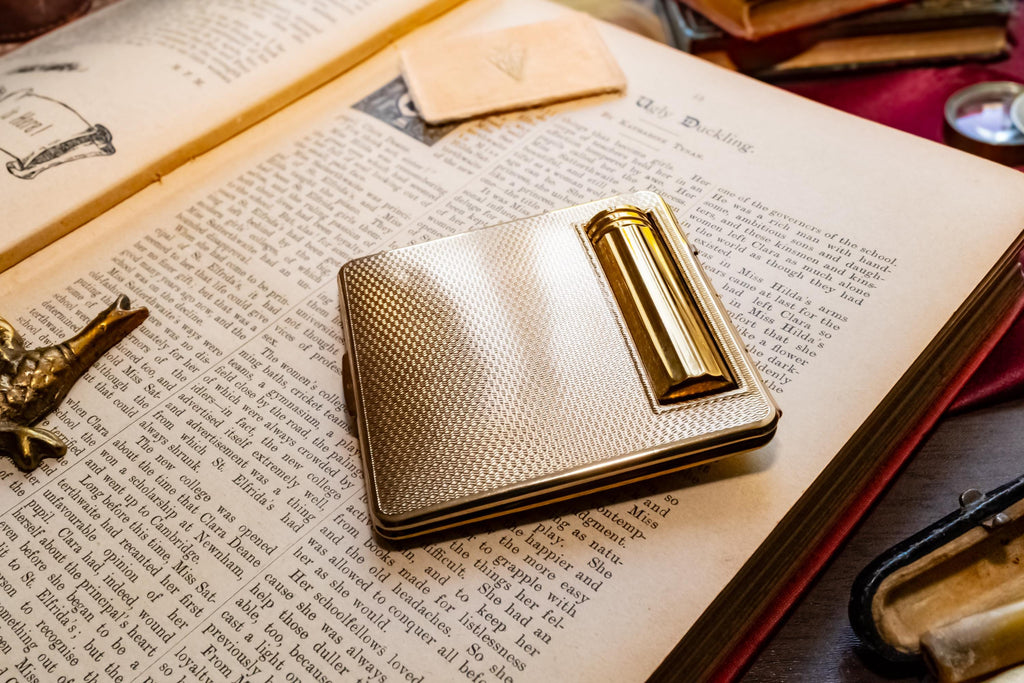 A gleaming gold vintage Kigu of London rectangular compact with a basketweave pattern and integrated lipstick holder, resting on an open antique book.