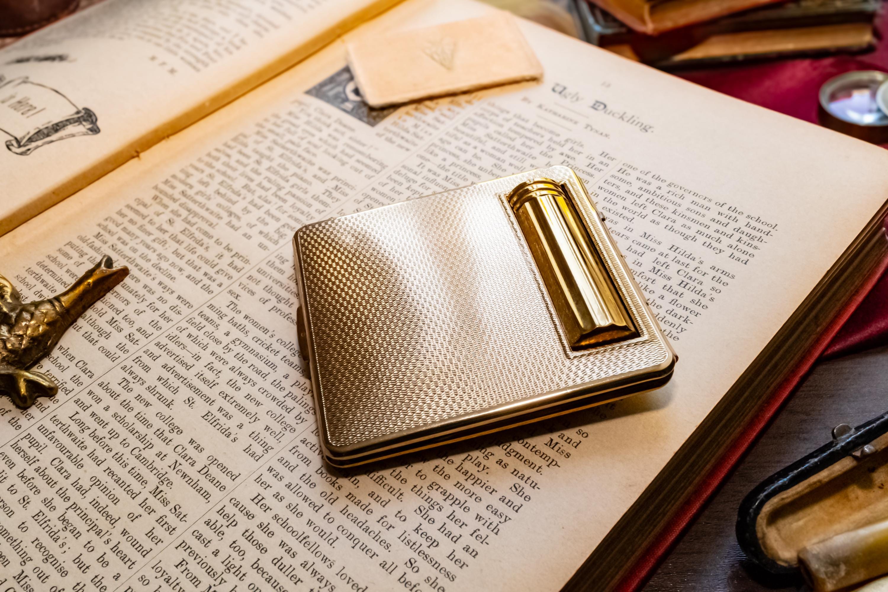 A gleaming gold vintage Kigu of London rectangular compact with a basketweave pattern and integrated lipstick holder, resting on an open antique book.