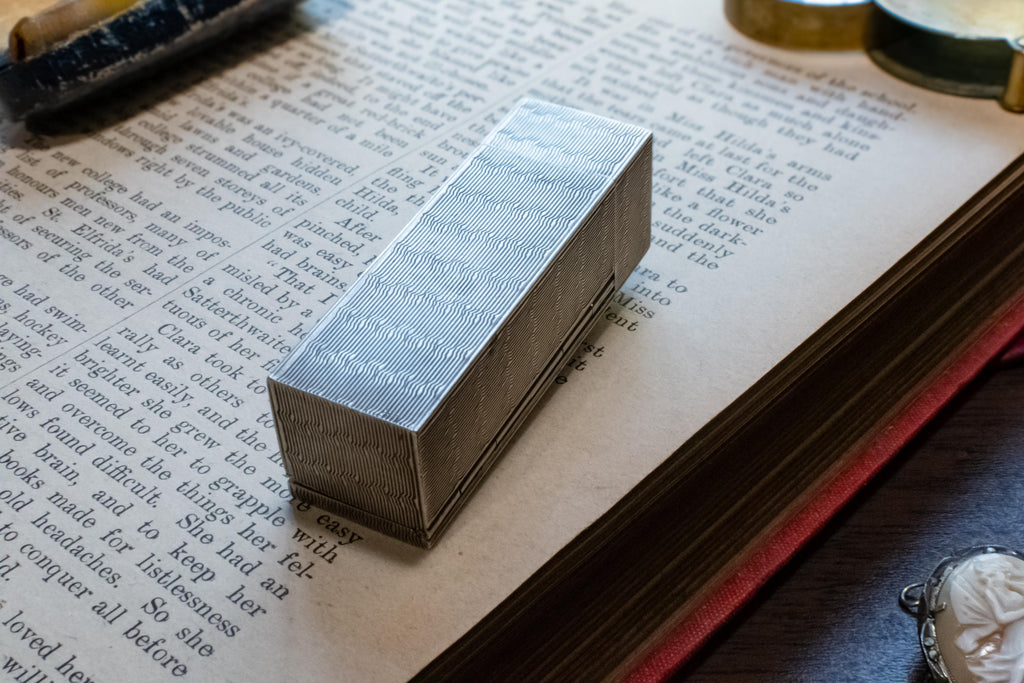 A rectangular silver metal box with engraved lettering, resting on the pages of an open vintage book.