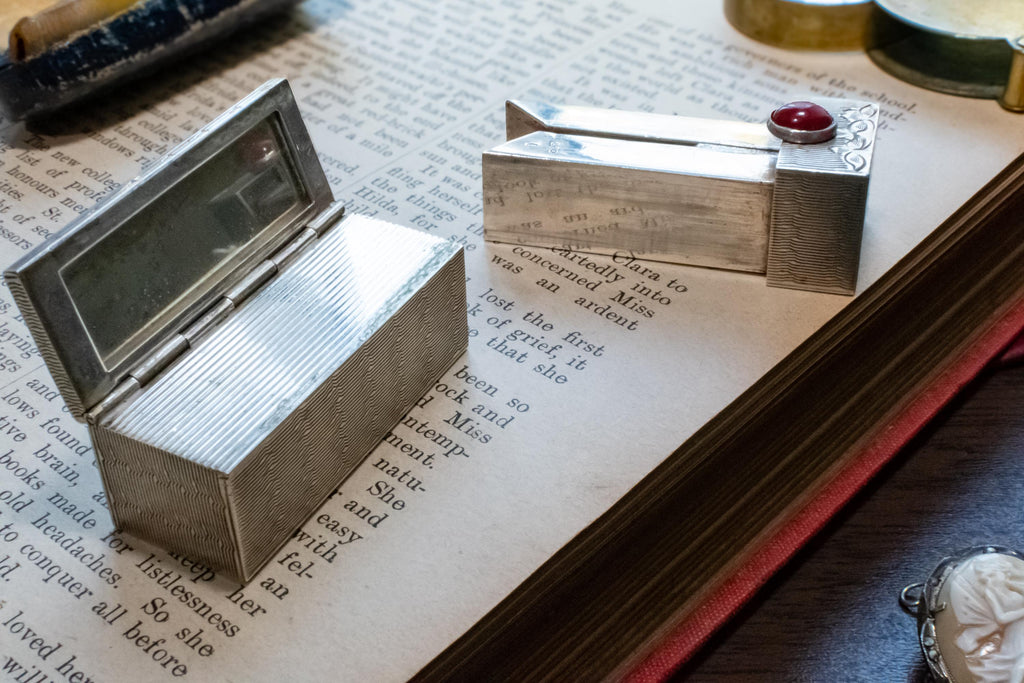 Two vintage silver-toned lipstick holder with mirrors and a red gemstone, displayed on an open antique book.