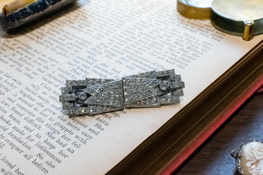 A silver-toned Art Deco brooch with intricate crystal or diamond inlay, resting on an open vintage book.