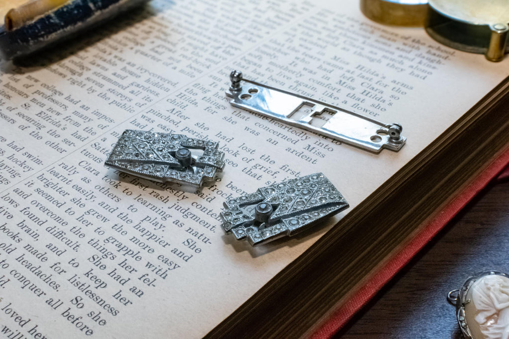 Two silver-toned ornate brooch displayed on an open book, alongside a metal watch spring component, with vintage props around them.