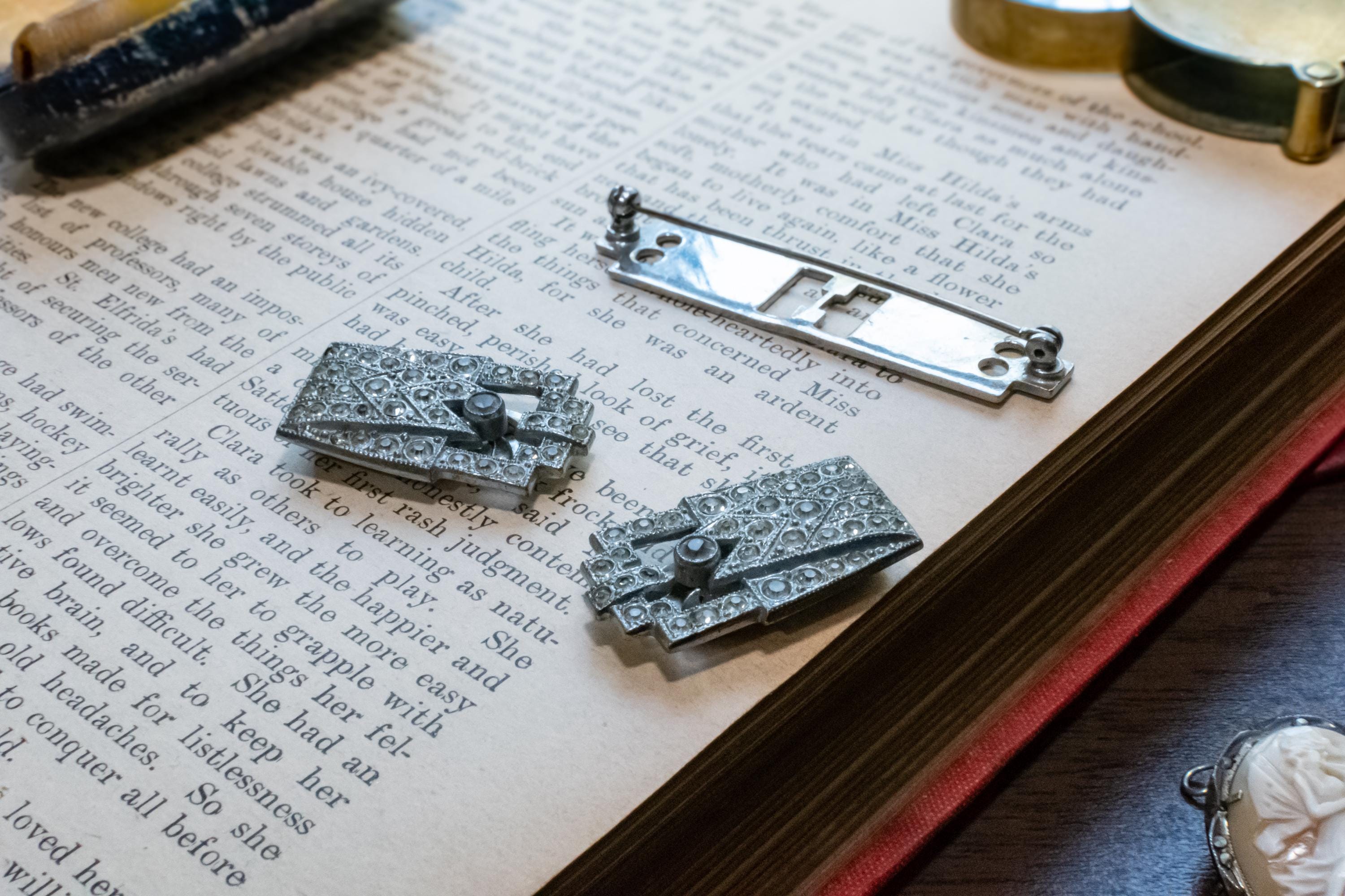 Two silver-toned ornate brooch displayed on an open book, alongside a metal watch spring component, with vintage props around them.