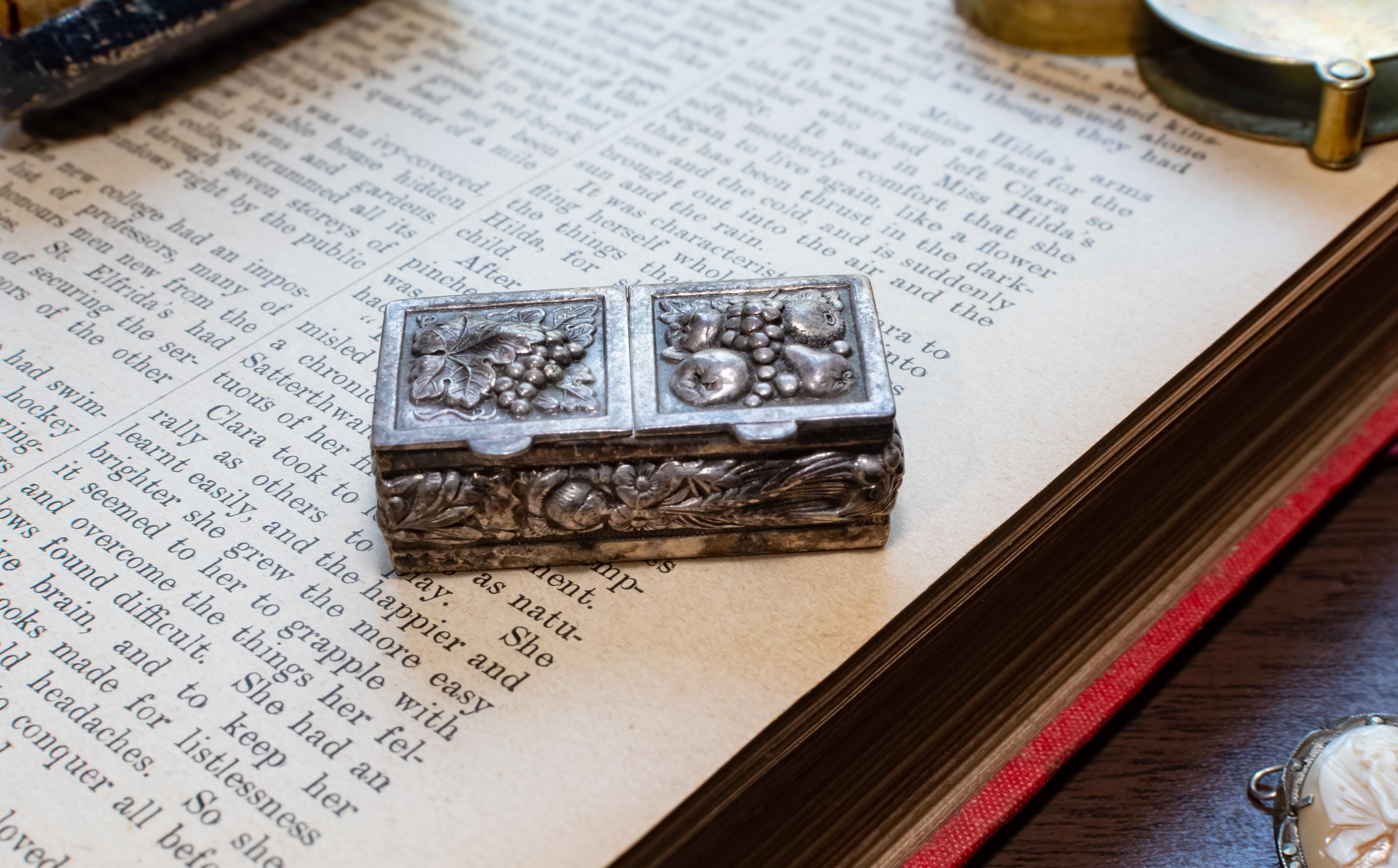 An ornate silver-toned metal keepsake box with floral and fruit motifs, resting on the pages of an open vintage book.
