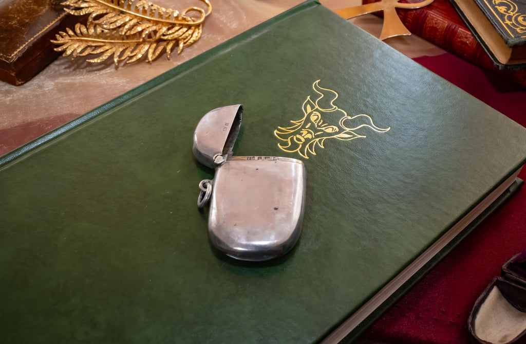Antique 1906 Edwardian sterling silver vesta case by Thomas Bishton, shown closed and open on a vintage book next to a brass magnifying glass.