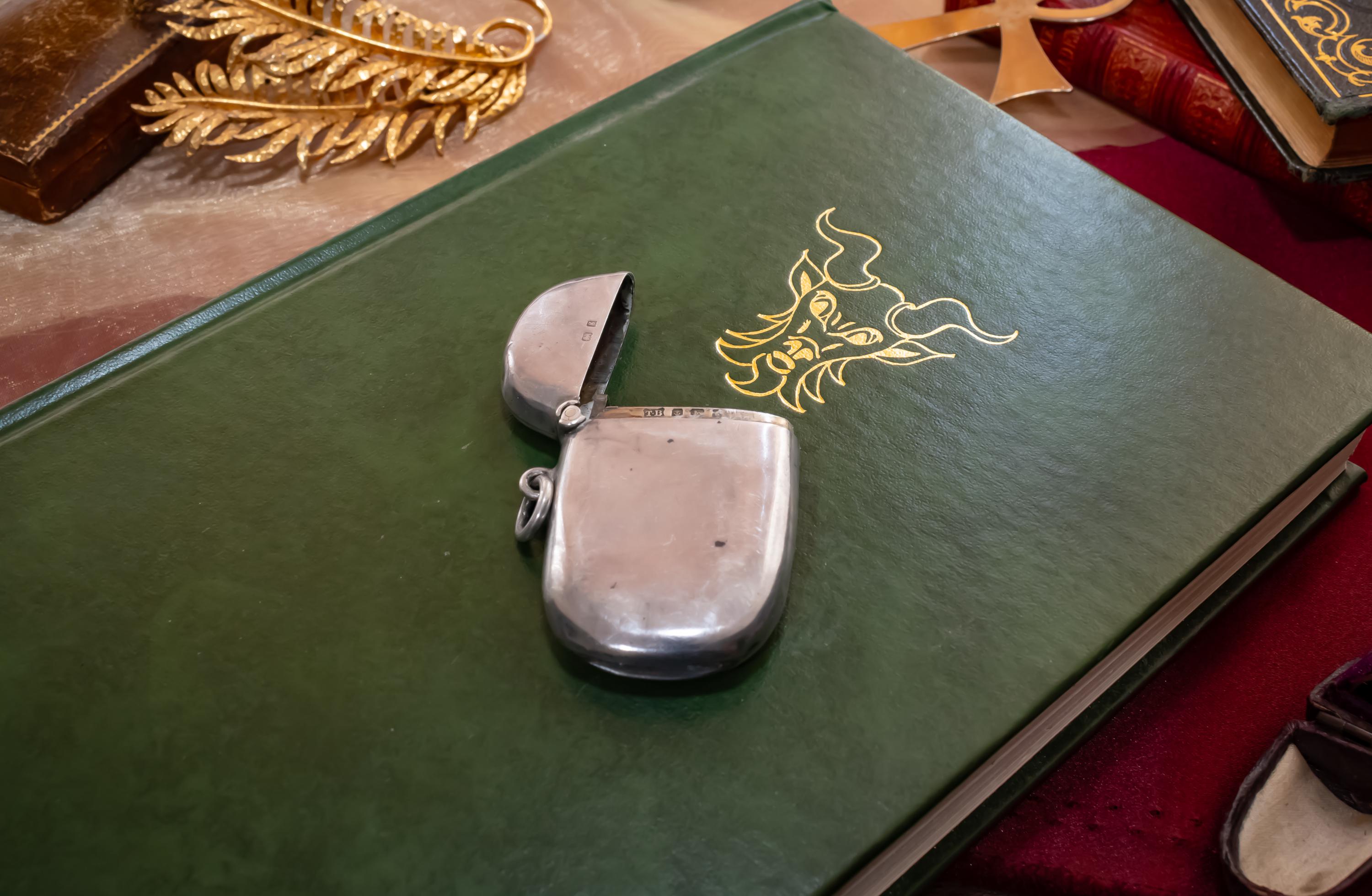 Antique 1906 Edwardian sterling silver vesta case by Thomas Bishton, shown closed and open on a vintage book next to a brass magnifying glass.