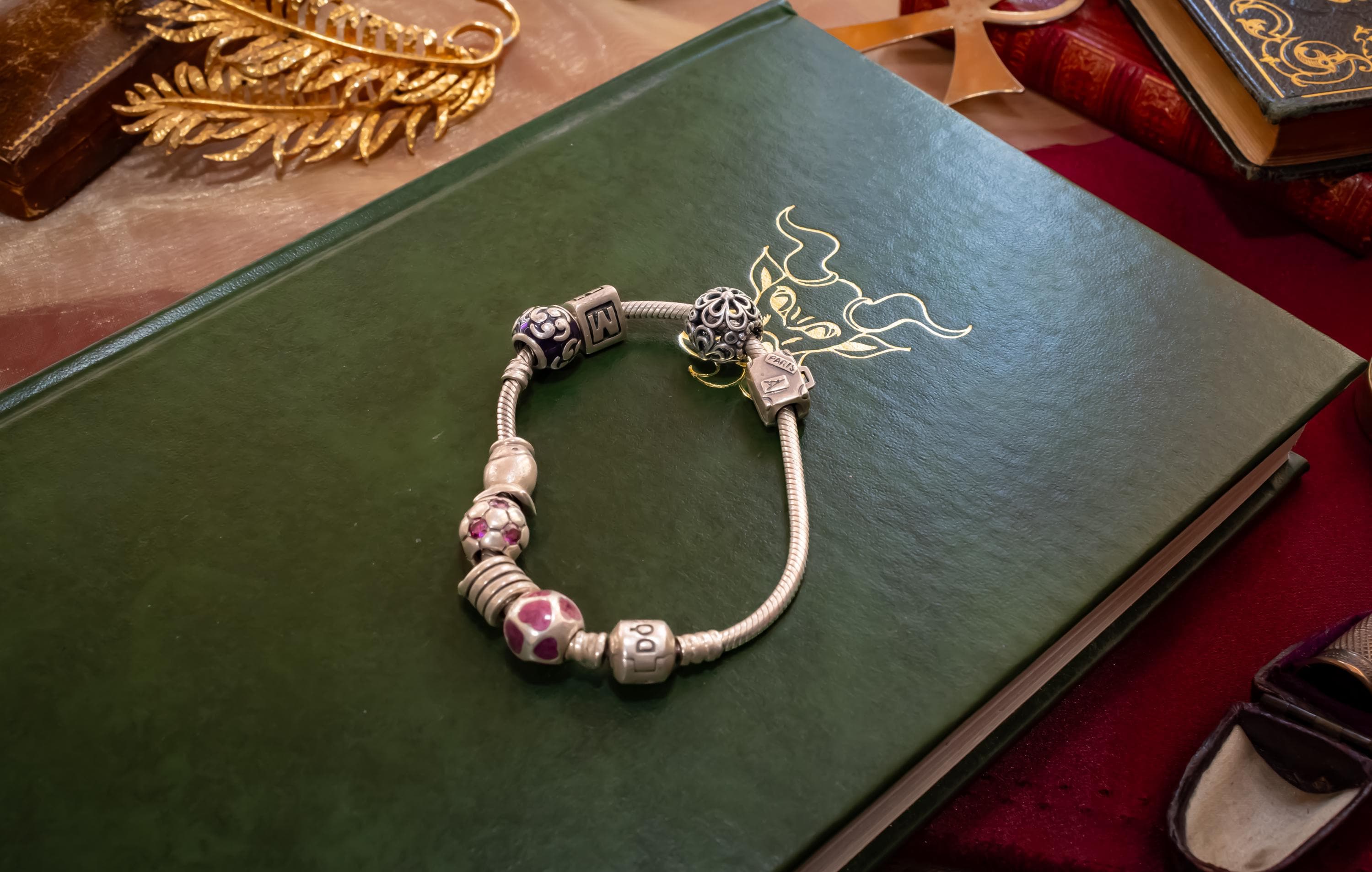 Authentic Pandora sterling silver snake chain bracelet loaded with nine various charms including a letter M and purple enamel beads, resting on an open antique book.