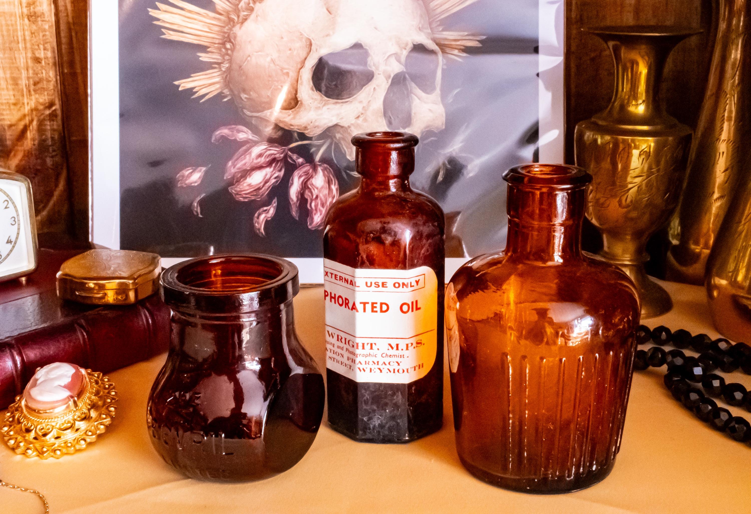 Three vintage amber glass apothecary bottles in varying shapes and sizes, one retaining a vintage label, displayed near a brass vase and skull artwork.