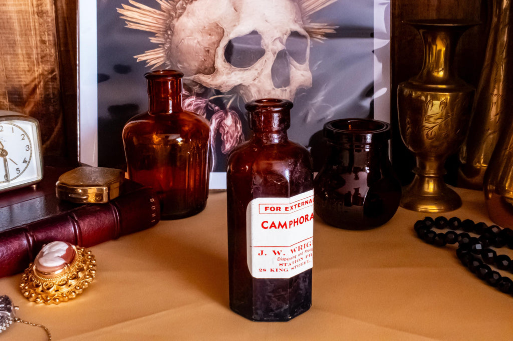 Three vintage amber glass apothecary bottles in varying shapes and sizes, one retaining a vintage label, displayed near a brass vase and skull artwork.