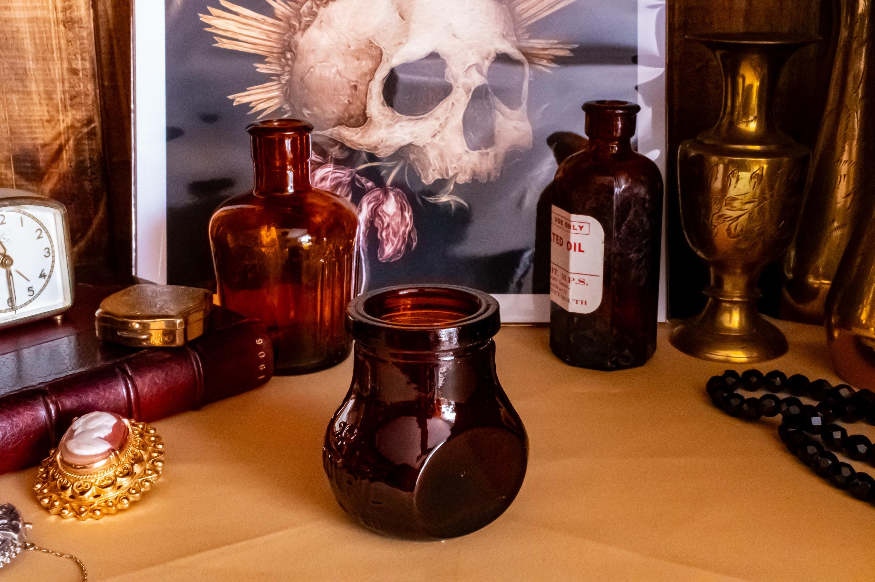 Three vintage amber glass apothecary bottles in varying shapes and sizes, one retaining a vintage label, displayed near a brass vase and skull artwork.