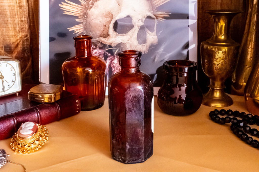 Three vintage amber glass apothecary bottles in varying shapes and sizes, one retaining a vintage label, displayed near a brass vase and skull artwork.