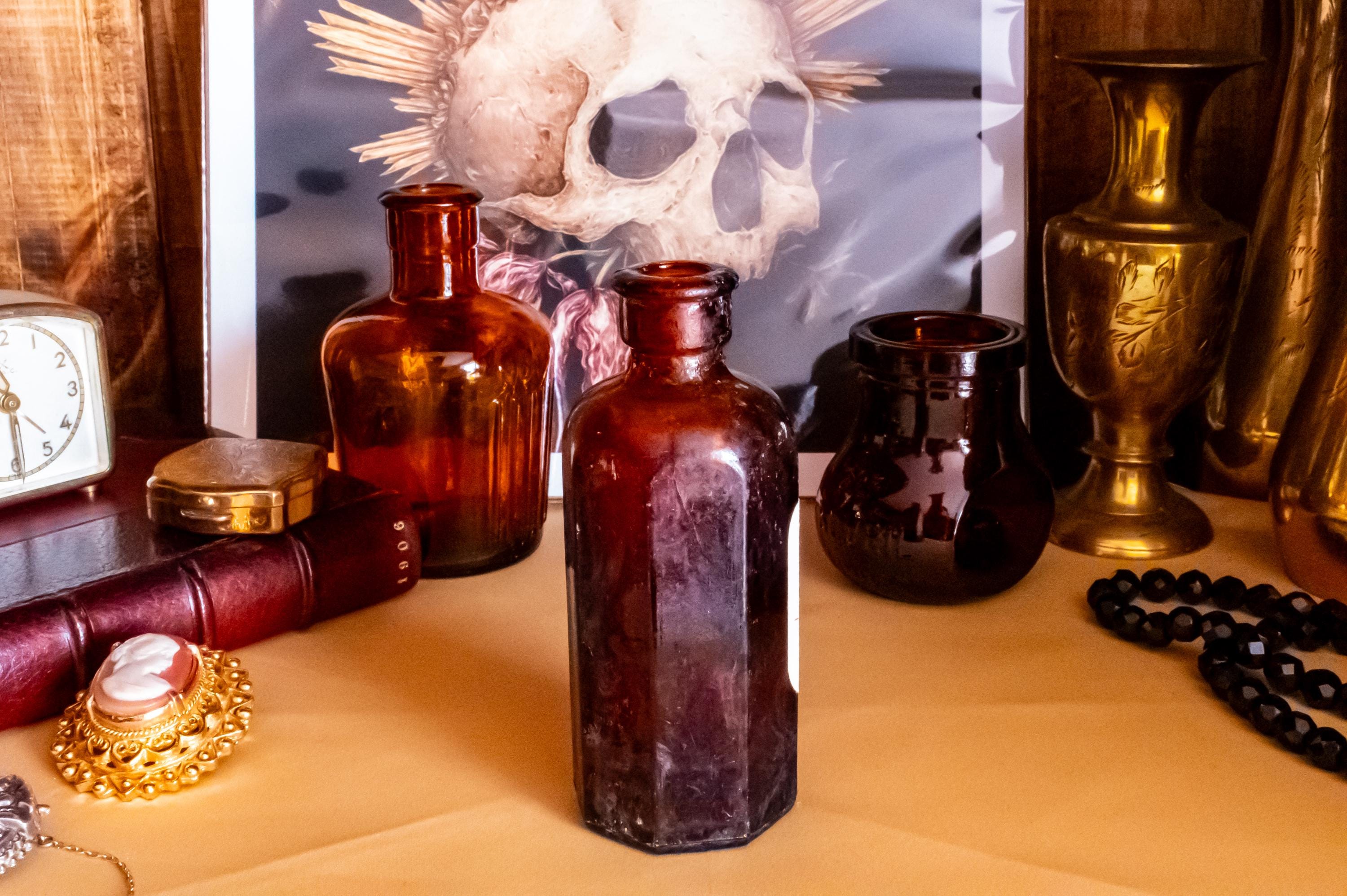 Three vintage amber glass apothecary bottles in varying shapes and sizes, one retaining a vintage label, displayed near a brass vase and skull artwork.