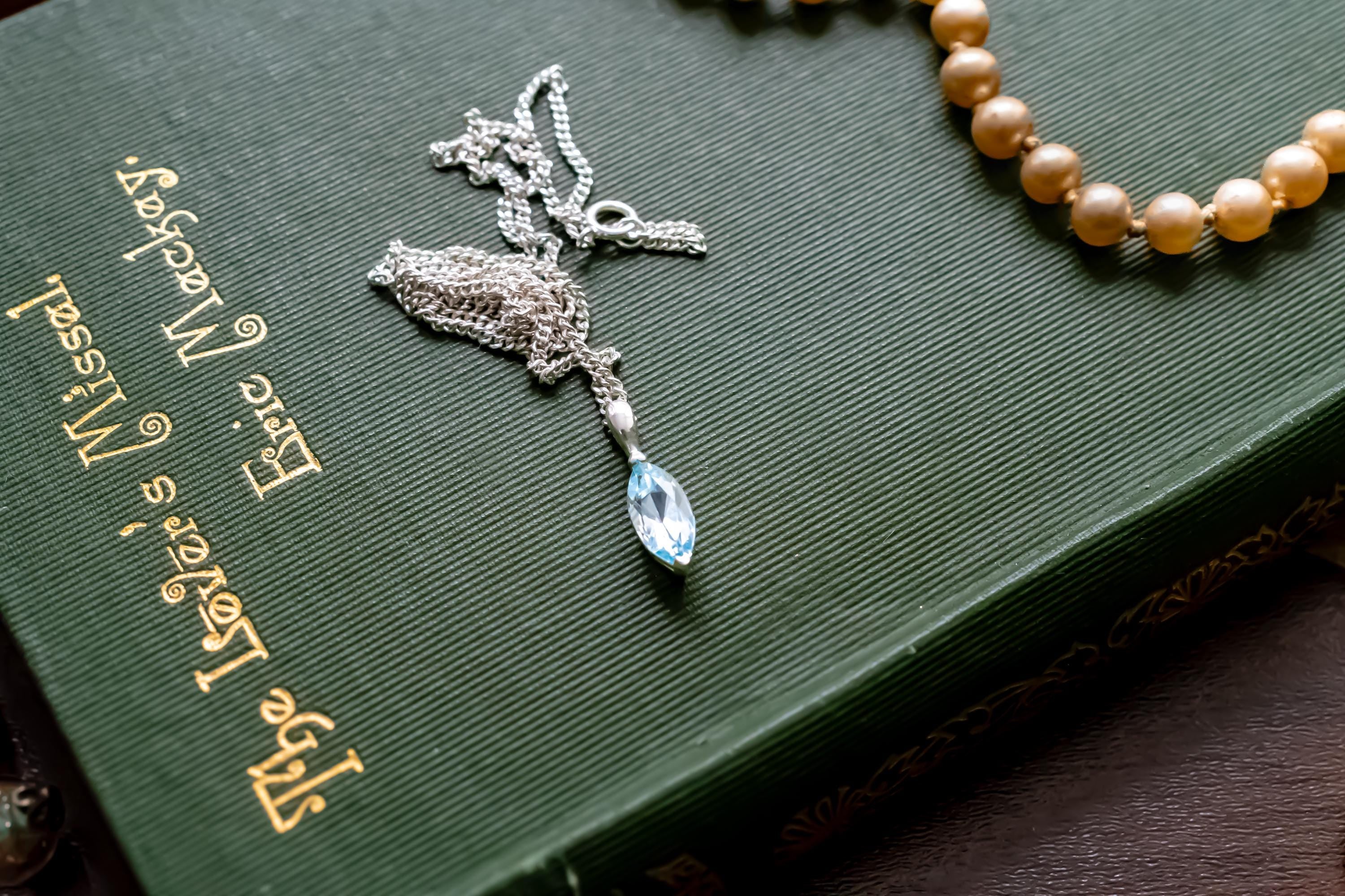 Delicate vintage 925 sterling silver necklace featuring a faceted teardrop-shaped light blue topaz gemstone, draped over a green antique book.