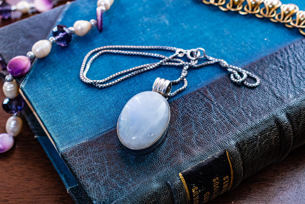 Large oval cabochon luminous moonstone pendant set in 925 sterling silver on a heavy box chain, displayed on a blue vintage book.